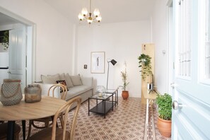 Una sala de estar acogedora con un sofá, una mesa de centro y sillas. Se aprecia una araña, un cuadro enmarcado y una planta en maceta.