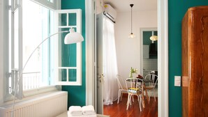 A room with a wooden floor, a white radiator, a window with white shutters, a white lamp, and a table with chairs. - Live in Athens, Thiseio Historic House (Athens)