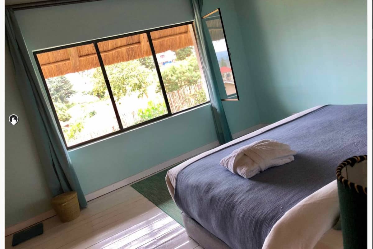 A bedroom with a bed, a window with a view of greenery, and a small trash bin.