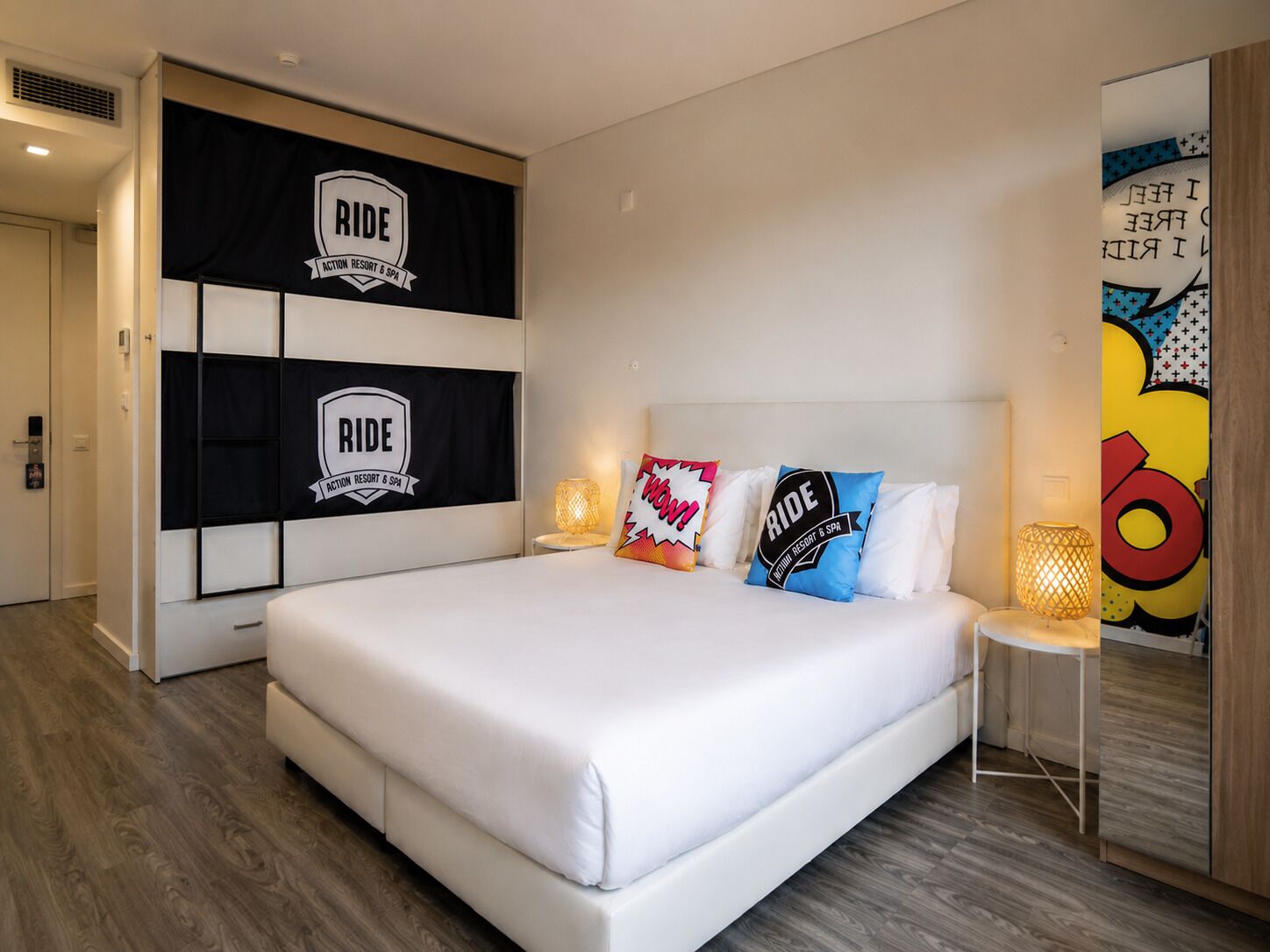 A modern hotel room with a large bed, a wall-mounted TV, and a decorative pillow with 'RIDE' branding.