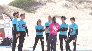 A group of people in wetsuits on a sandy beach with surfboards, some with 'SURF ACADEMY' and 'RIDE' logos.