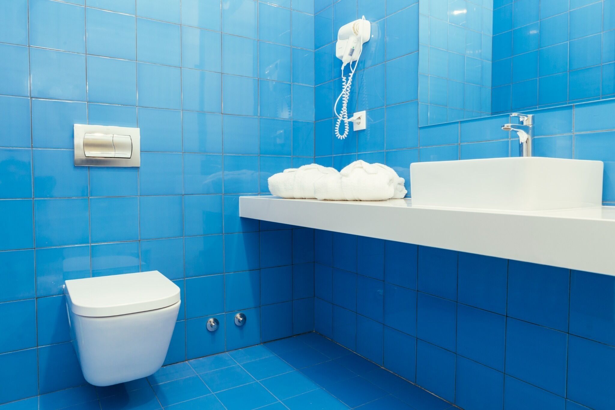 A bathroom with blue tiles, a white toilet, a sink, and a hairdryer on the wall.