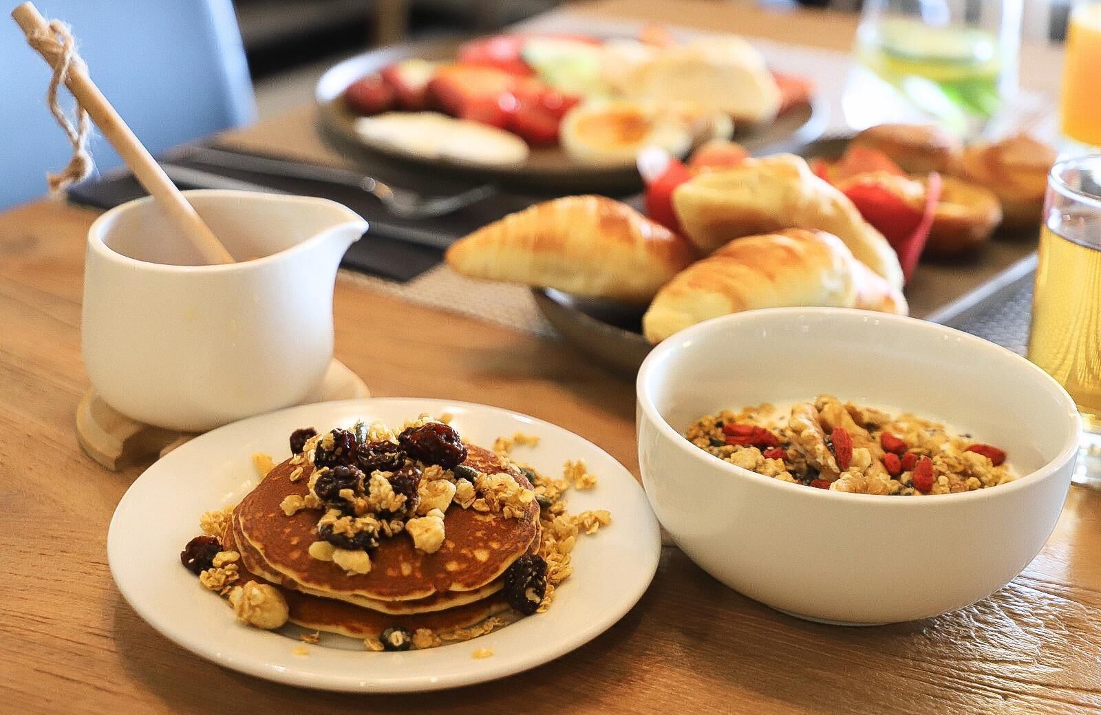 A breakfast setting with pancakes topped with nuts and dried fruits, a bowl of granola, and croissants on a wooden table.