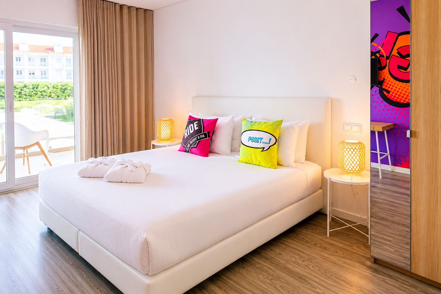 Ein Hotelzimmer mit einem großen Bett, einem Fenster mit Vorhängen, einem Nachttisch mit Lampe und Blick auf Gebäude.