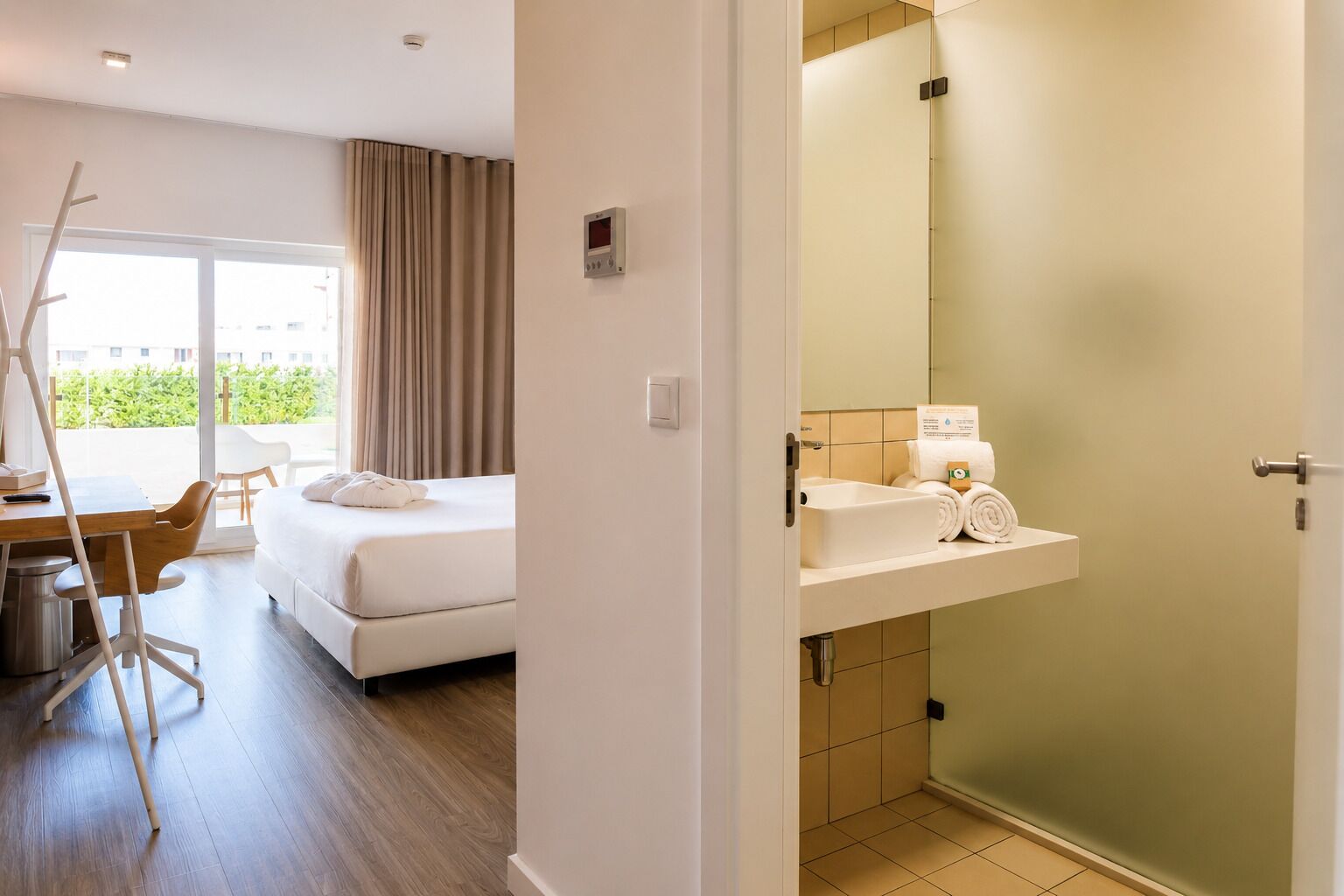 A hotel room with a bed, a desk, a chair, a bathroom with a sink and a mirror, and a view of greenery outside.