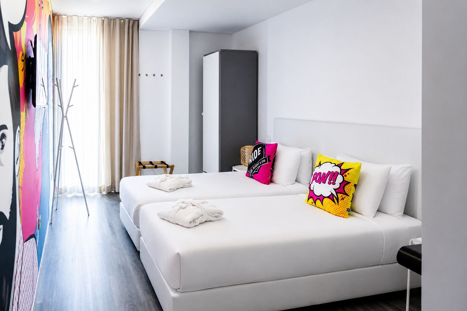 A modern hotel room with two beds, a large window with curtains, a wardrobe, and a colorful artwork on the wall.