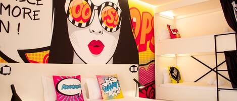 A modern hotel room with a large poster of a woman wearing glasses and a bold text saying "NEED PRACTICE MORE". There are bunk beds with pillows and a lamp on the side table.