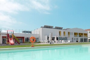 A swimming pool with a slide and sun loungers, surrounded by a white building with yellow accents and a lifebuoy in the pool.