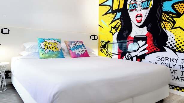 Una cama con ropa blanca, dos almohadas con diseños estilo cómic y un mural de pared llamativo.