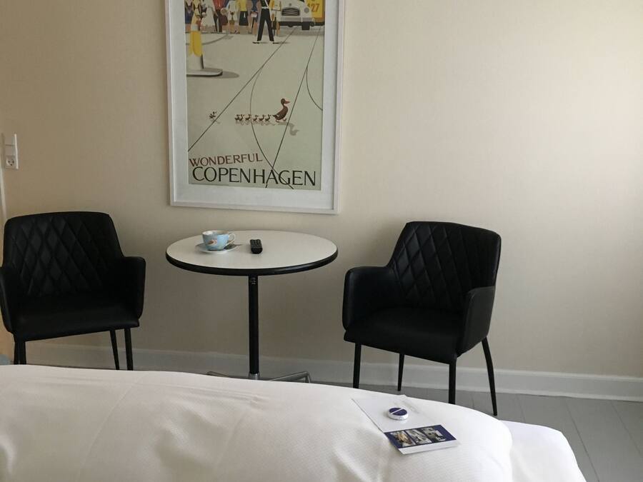 A hotel room with a bed, two black chairs, a small round table, and a framed poster on the wall.