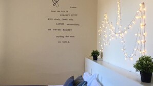 Un dormitorio con cama, almohadas, una pared con una cita motivacional, luces de cadena y dos plantas en macetas.