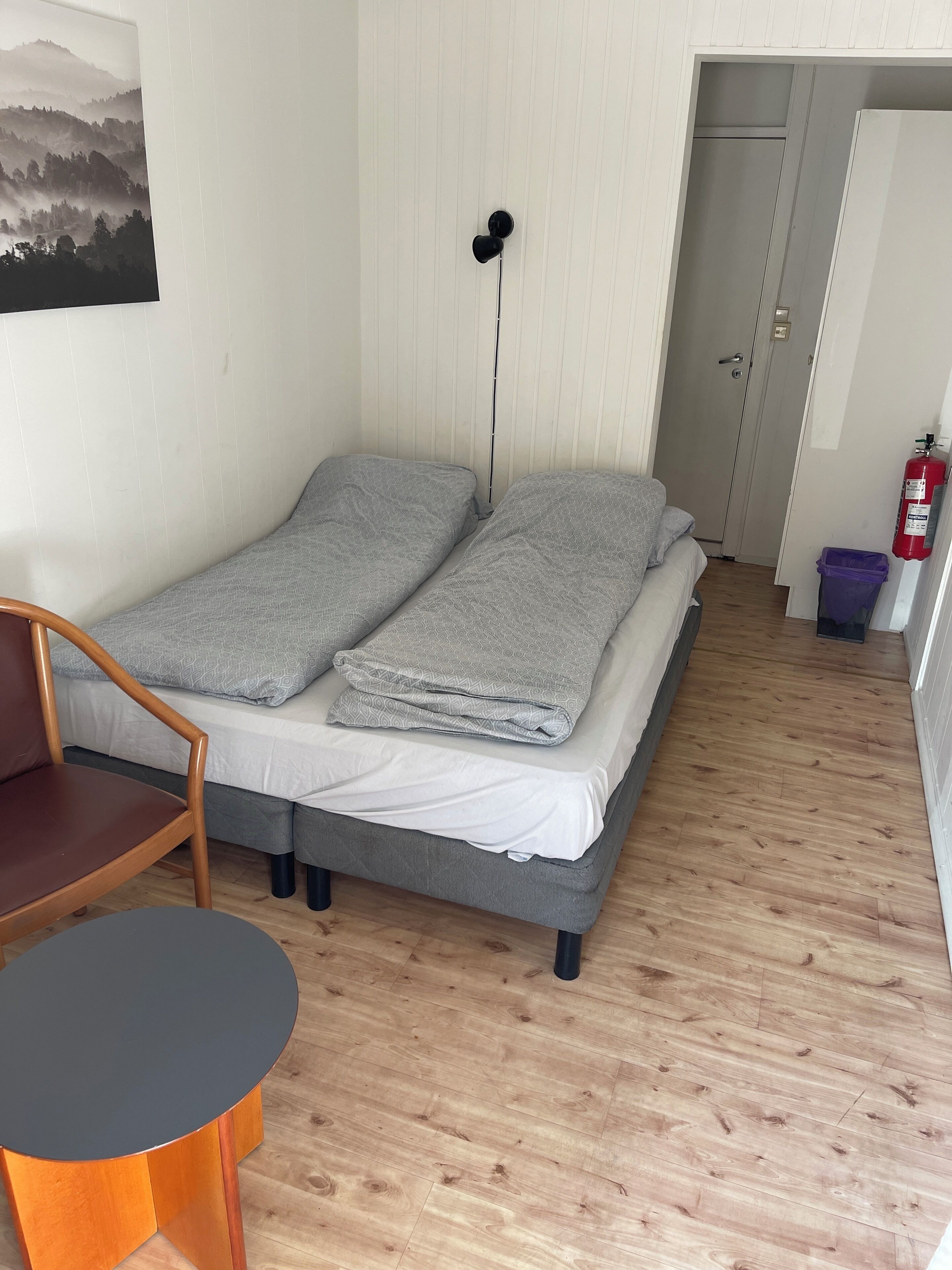 A small, single-bed room with a wooden floor, a chair, a table, and a door leading to another room.