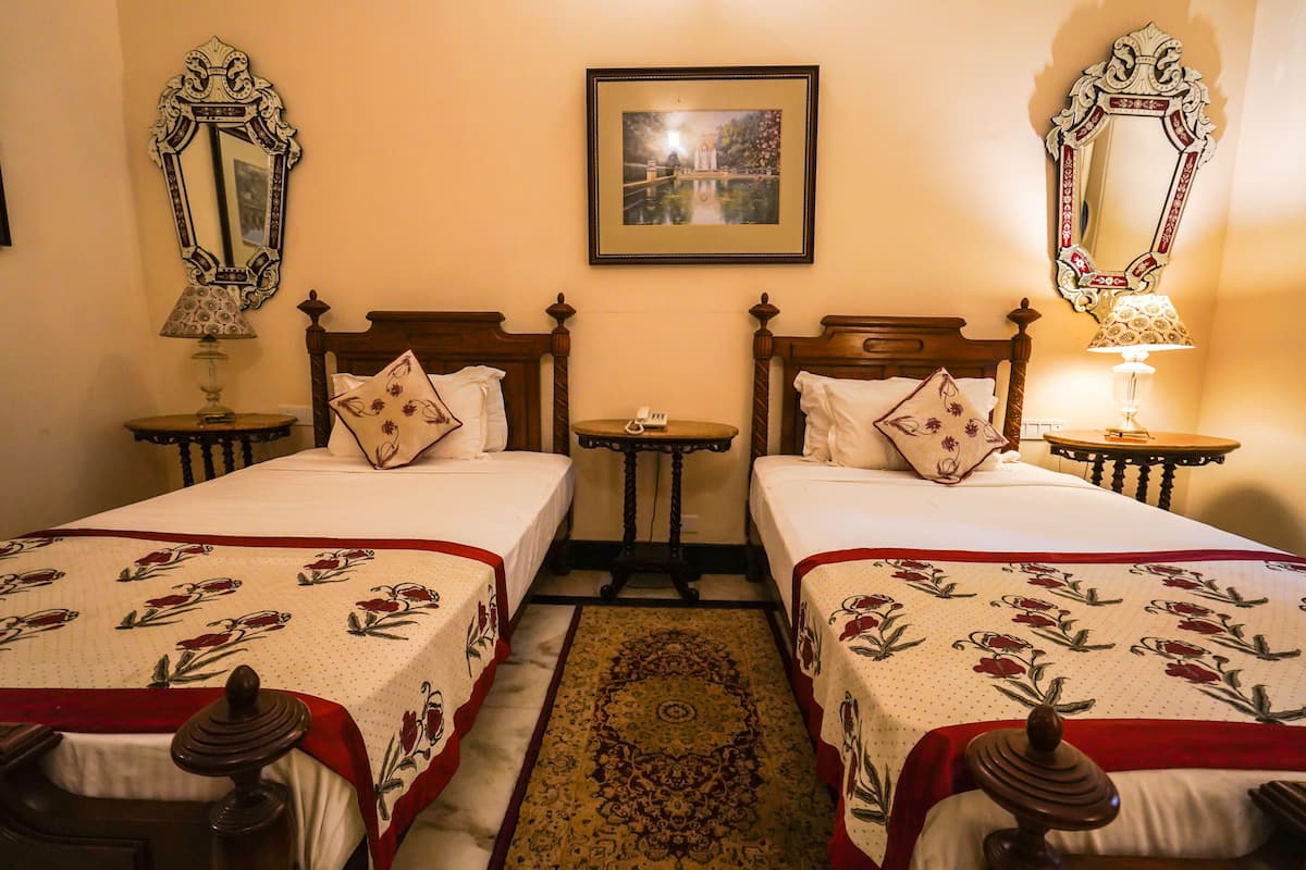 A hotel room with two single beds, a small table, a mirror, and a framed picture on the wall.