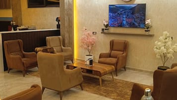 A hotel lobby with leather armchairs, a wooden coffee table, a TV mounted on the wall, and a reception desk.