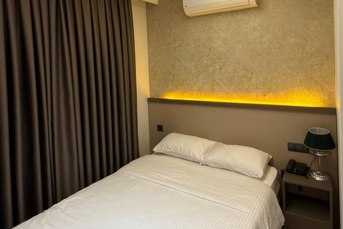 A modern hotel room with a bed, a nightstand, an air conditioning unit, and a ceiling light fixture.
