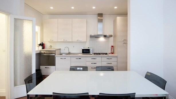 Une cuisine moderne avec des armoires blanches, une hotte de cuisinière en acier inoxydable, un four à micro-ondes et une cuisinière à gaz. On y trouve une table à manger avec des chaises et une cloison en verre.