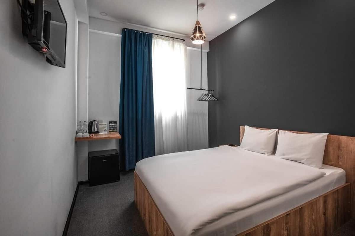A hotel room with a bed, a desk with a kettle, a mirror, and a window with curtains.