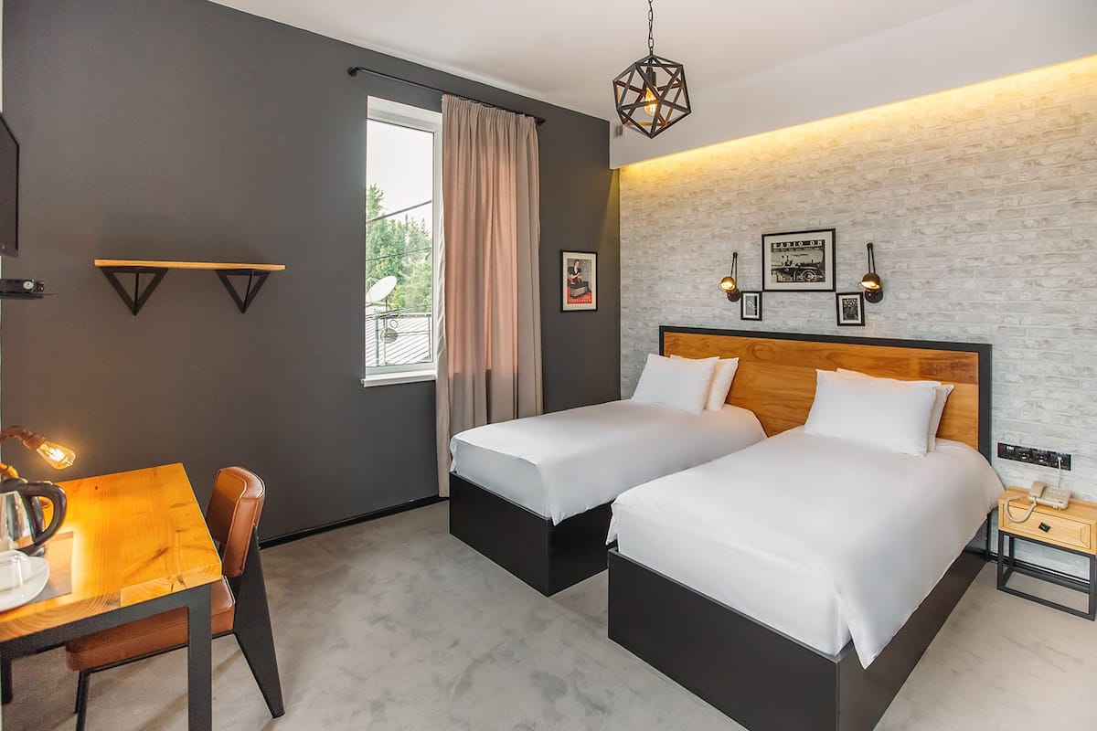 A hotel room with two beds, a wooden desk, a chair, a table, and a window with curtains.