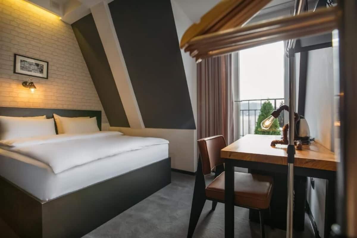 A hotel room with a bed, a desk, a chair, a window with a view, and a wall-mounted lamp.