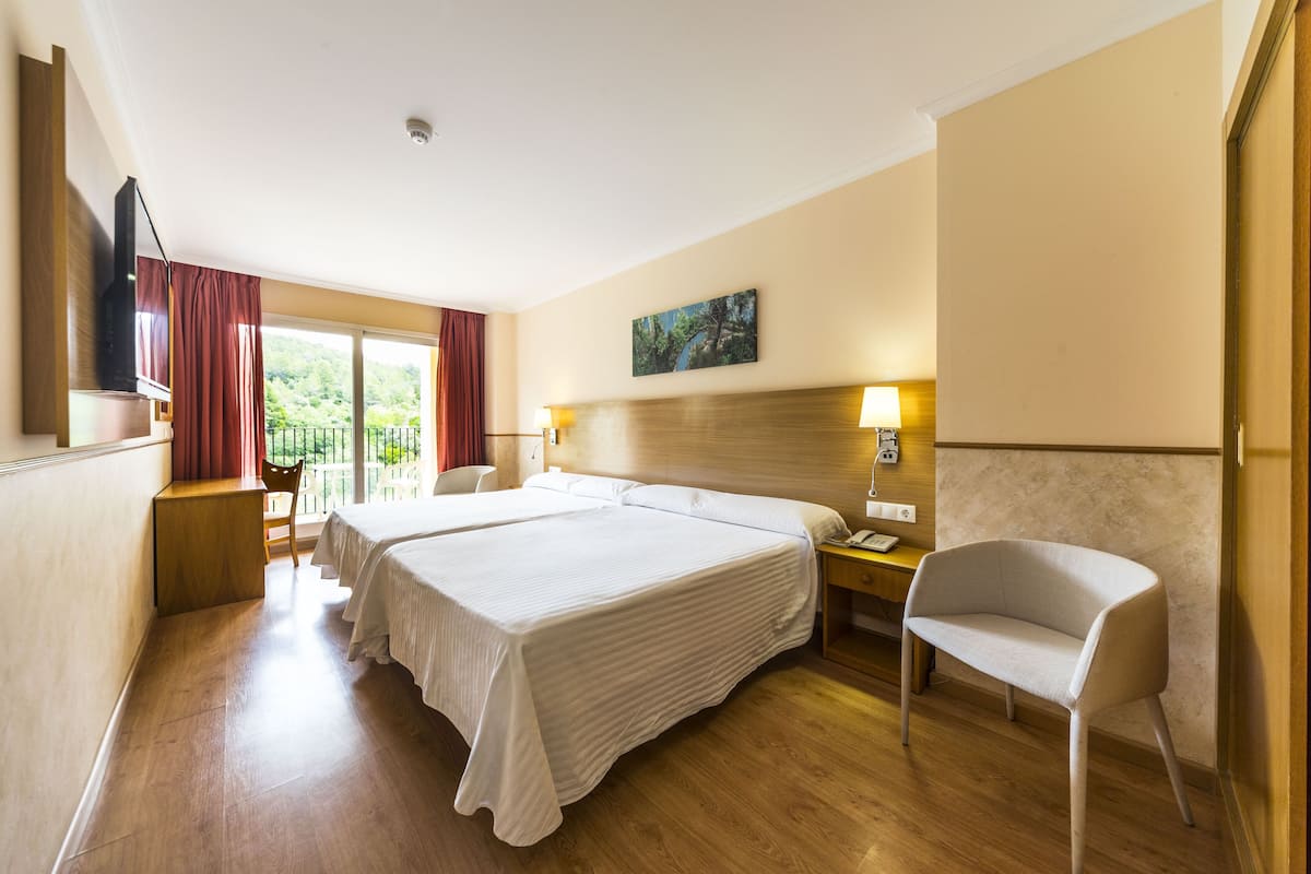 A hotel room with two beds, a wooden floor, a desk with a chair, a TV, and a window with curtains.