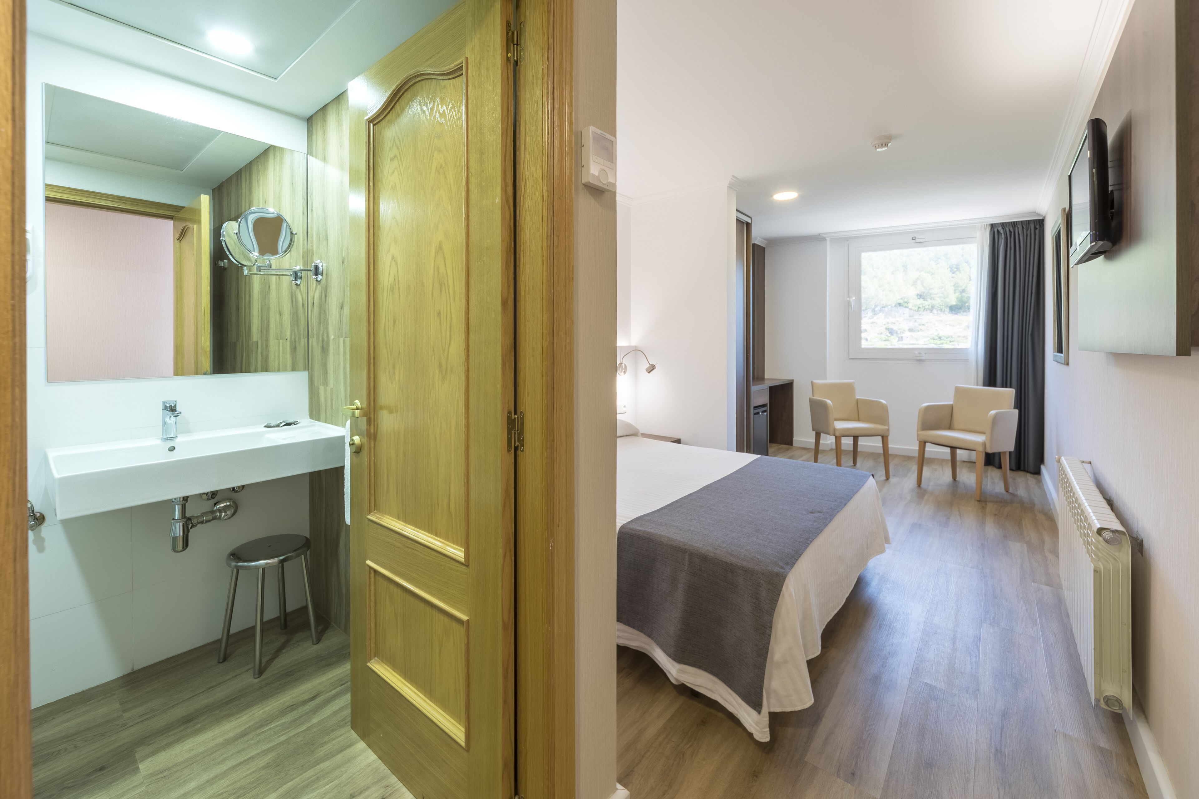 Habitación de hotel con puerta de madera, un lavabo blanco, un espejo, una cama con manta gris, dos sillas y un televisor en la pared.