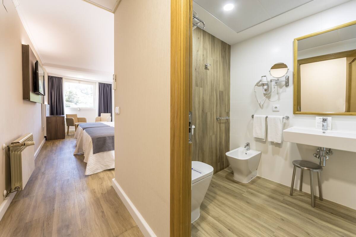 A hotel room with a wooden floor, a bed with a grey blanket, a bedside table, a TV, a window with curtains, a bathroom with a sink, mirror, and bidet.