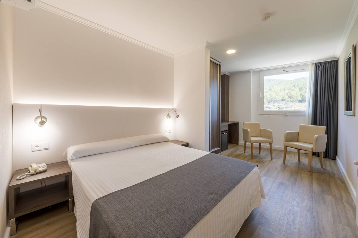 A hotel room with a large bed, bedside tables with lamps, a desk with chairs, and a window with curtains.
