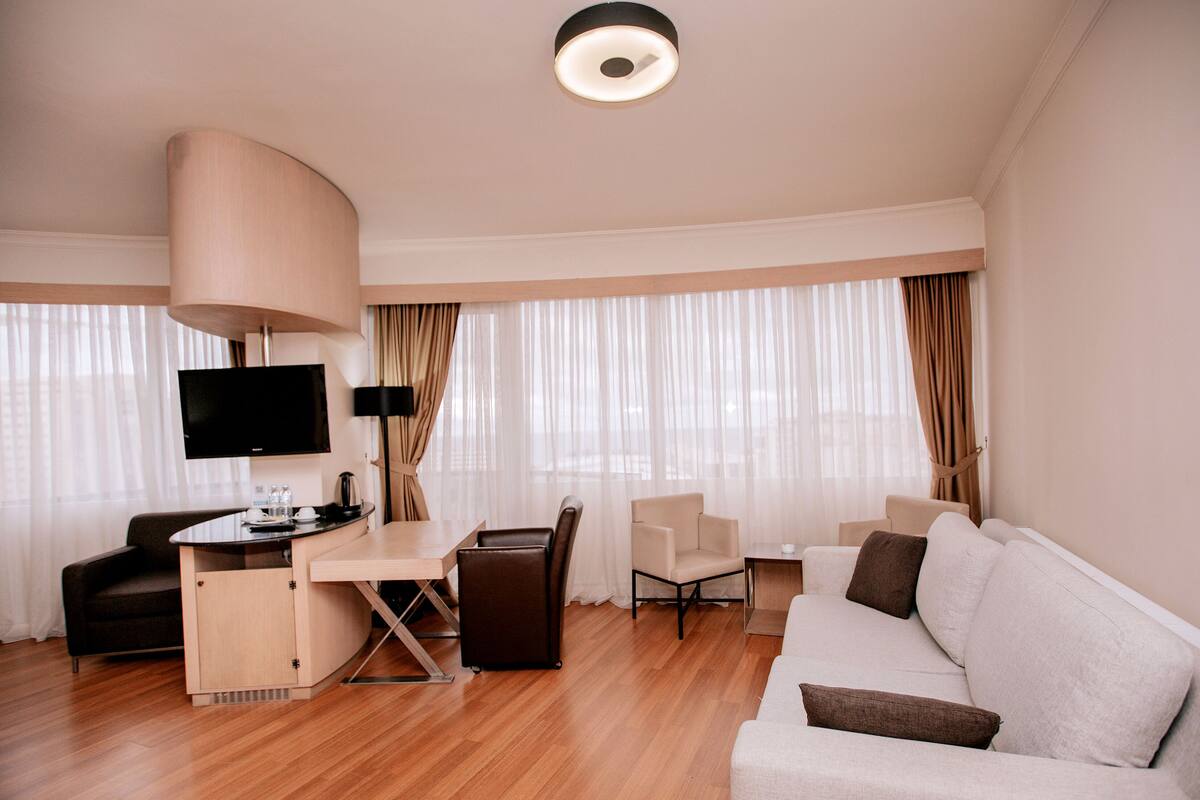 Una habitación de hotel moderna con suelo de madera, un sofá, un escritorio con una silla, un televisor y un ventanal con cortinas.