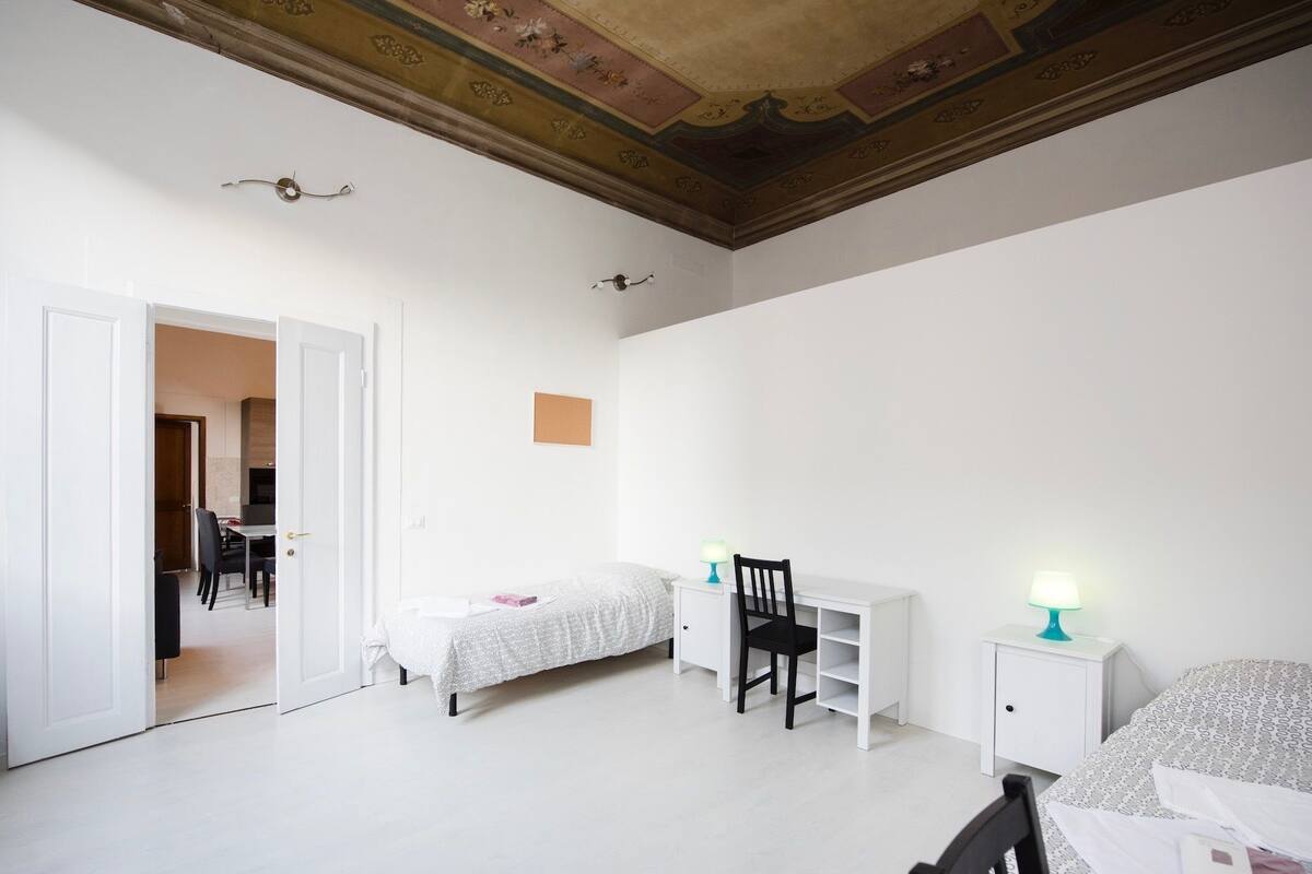 A bedroom with a bed, a desk, a chair, and a nightstand with a lamp.
