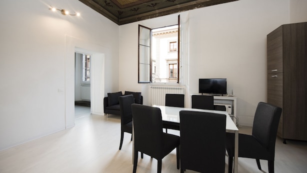 A dining area with a white table and black chairs, a TV on a cabinet, and a view of a building through a window. - Campidoglio (Florence)