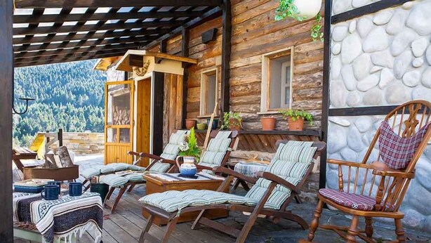 A wooden cabin with a porch, furnished with striped chairs and a rocking chair, overlooking a forested mountain.