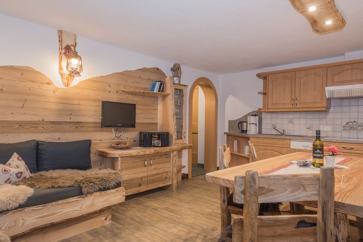 Un interior rústico con un sofá de madera, una mesa de comedor y una zona de cocina con armarios de madera y salpicadero de azulejos.