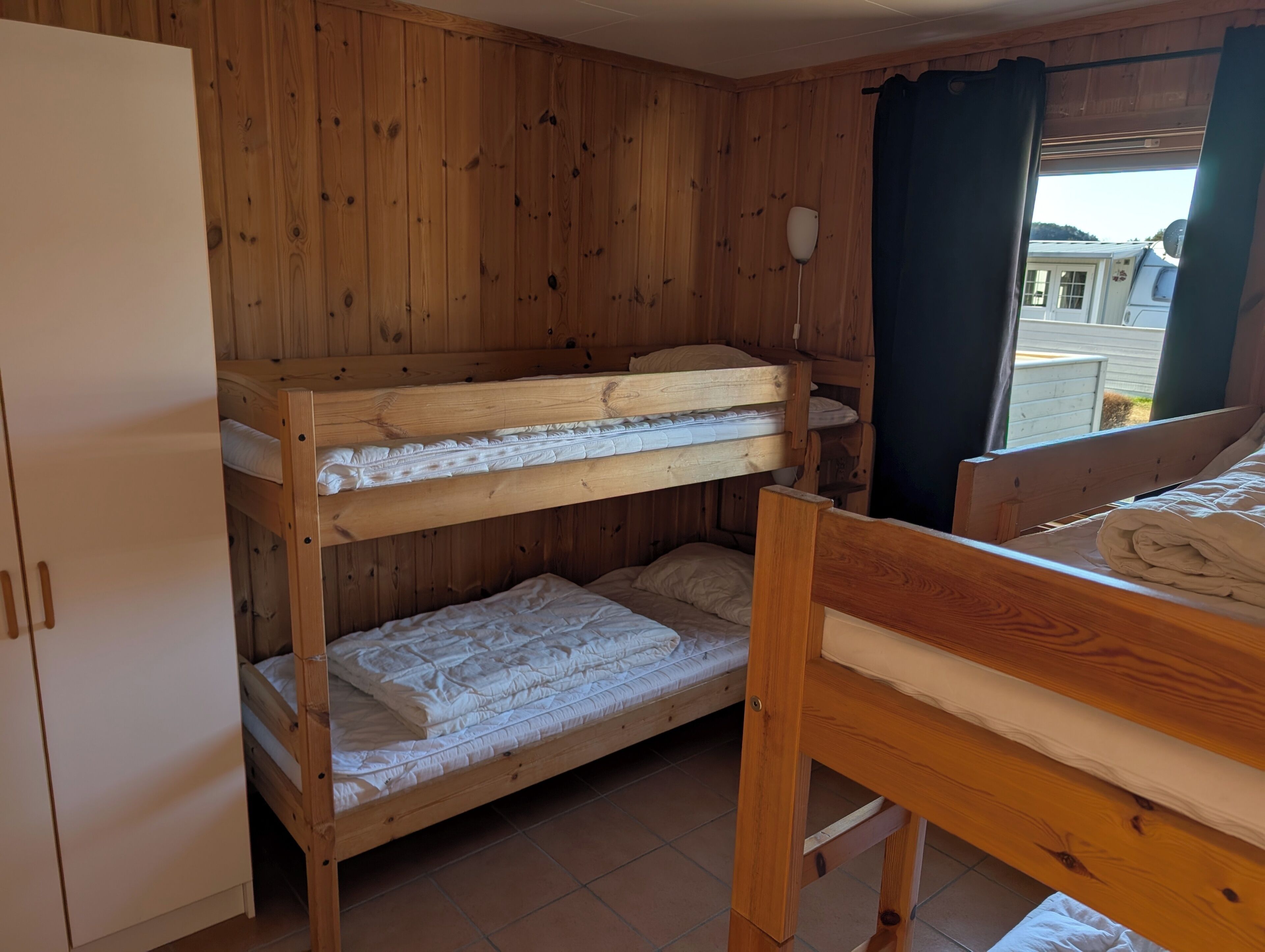 A bunk bed room with wooden walls, a white wardrobe, and a window with curtains.
