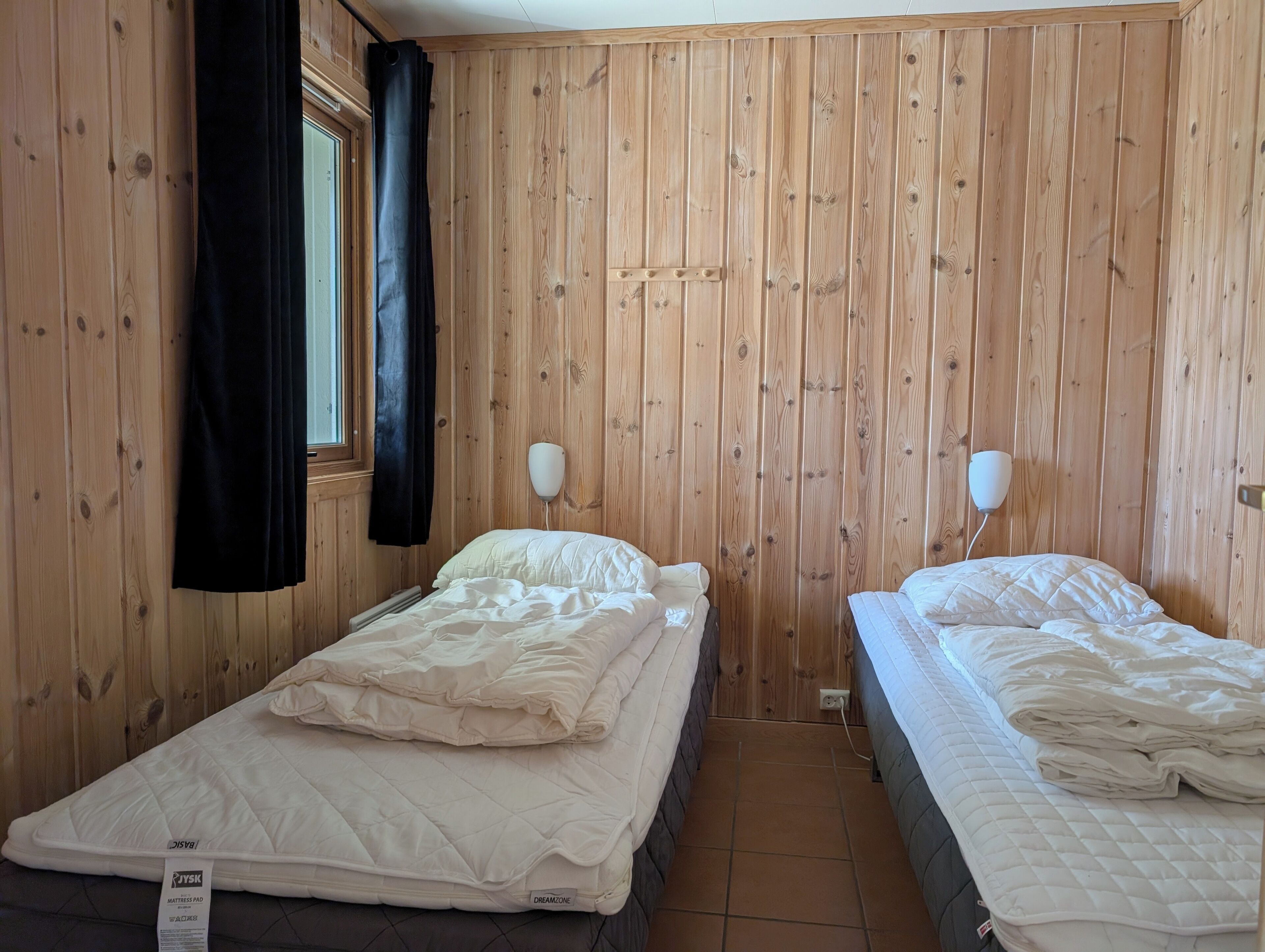 Ett rum med träpanel på väggarna, två sängar med vita sängkläder, ett fönster med mörka gardiner och en vägglampa.