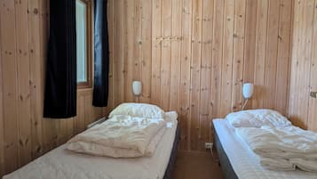 Ett rum med träpanel på väggarna, två sängar med vita sängkläder, ett fönster med mörka gardiner och en vägglampa.
