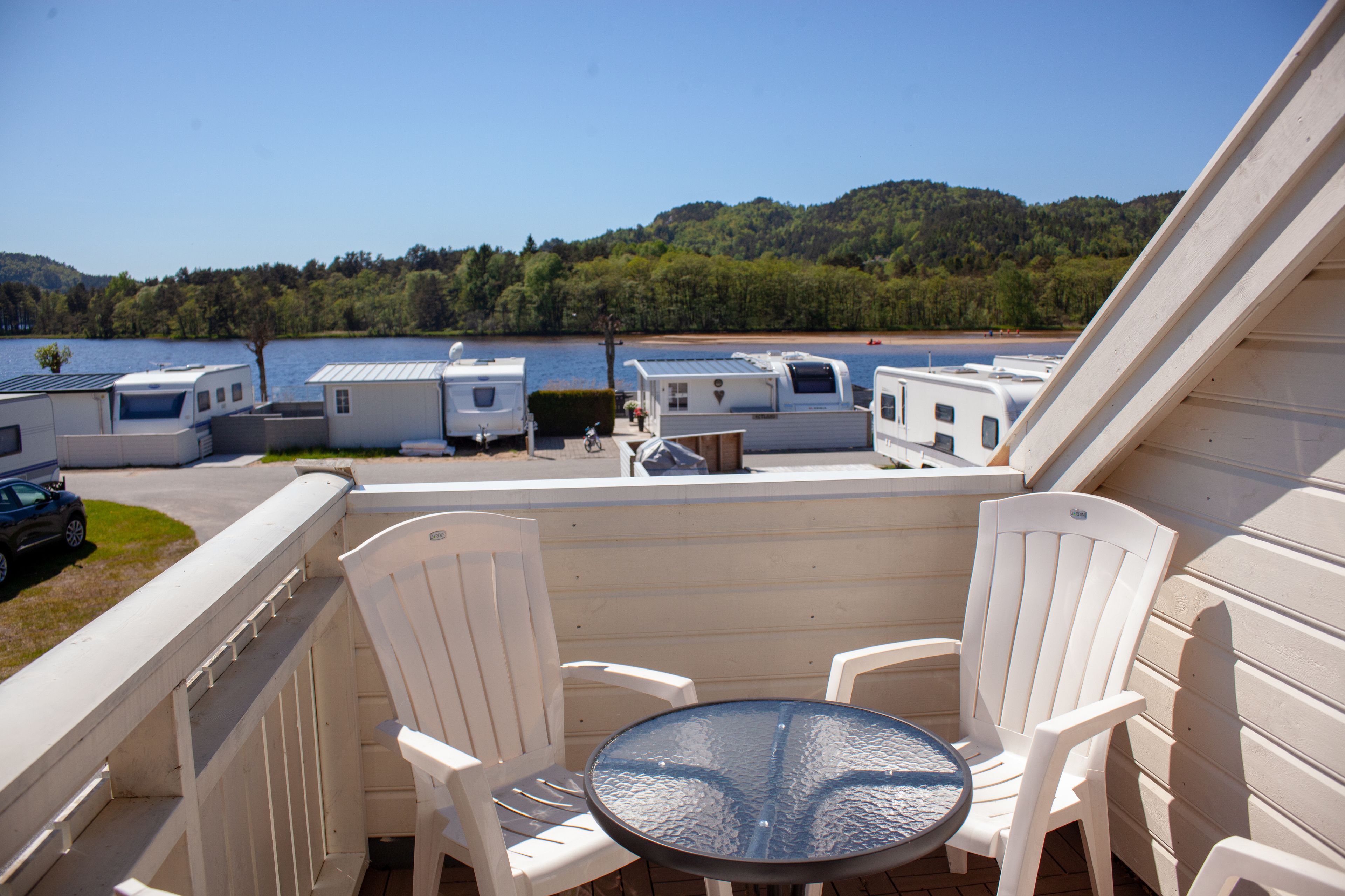A balcony with white chairs and a round table, overlooking a marina with RVs and a lake.