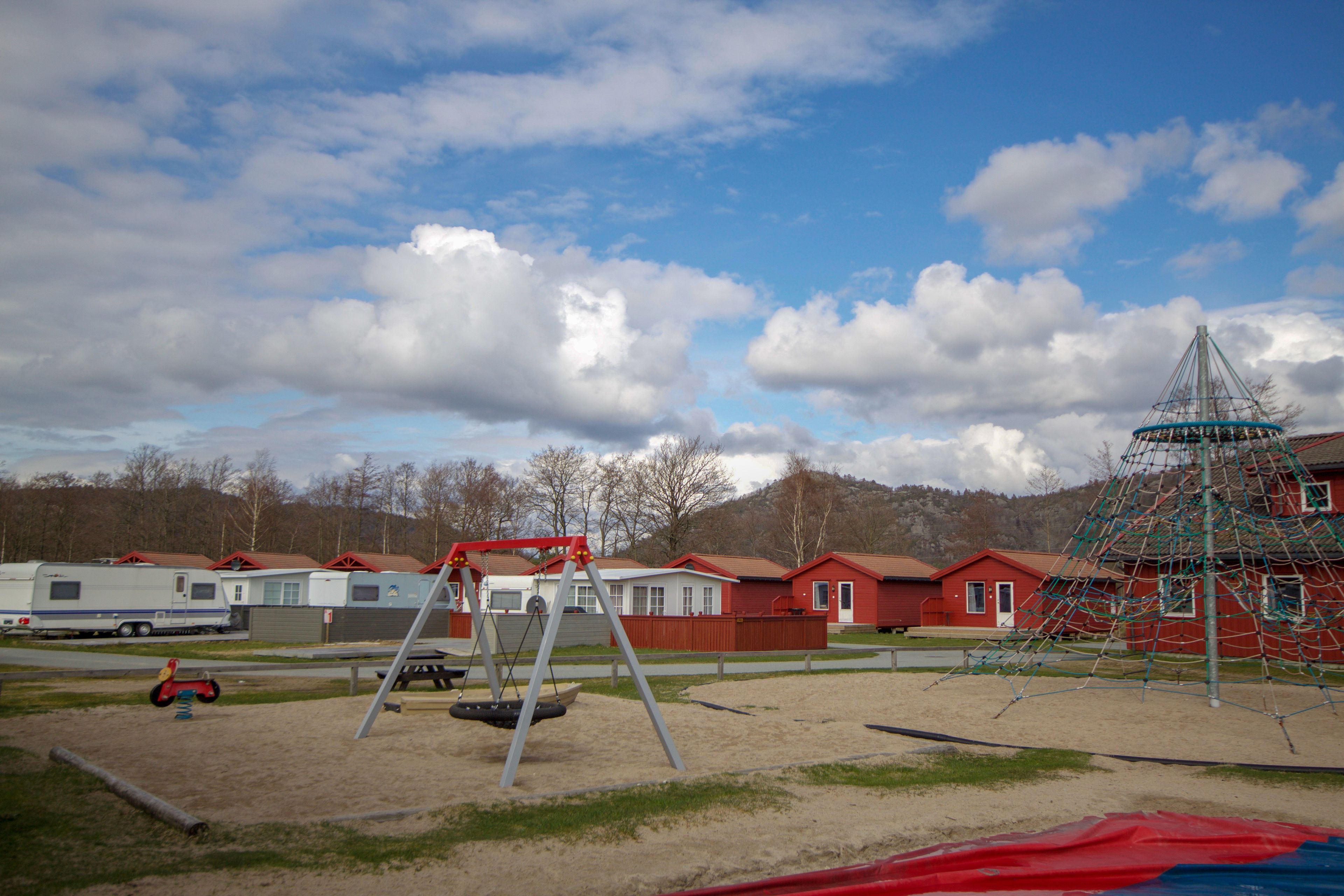 Ein Spielplatz mit Schaukeln und einer Kletterkonstruktion, umgeben von roten Häusern und einem Wohnwagen.