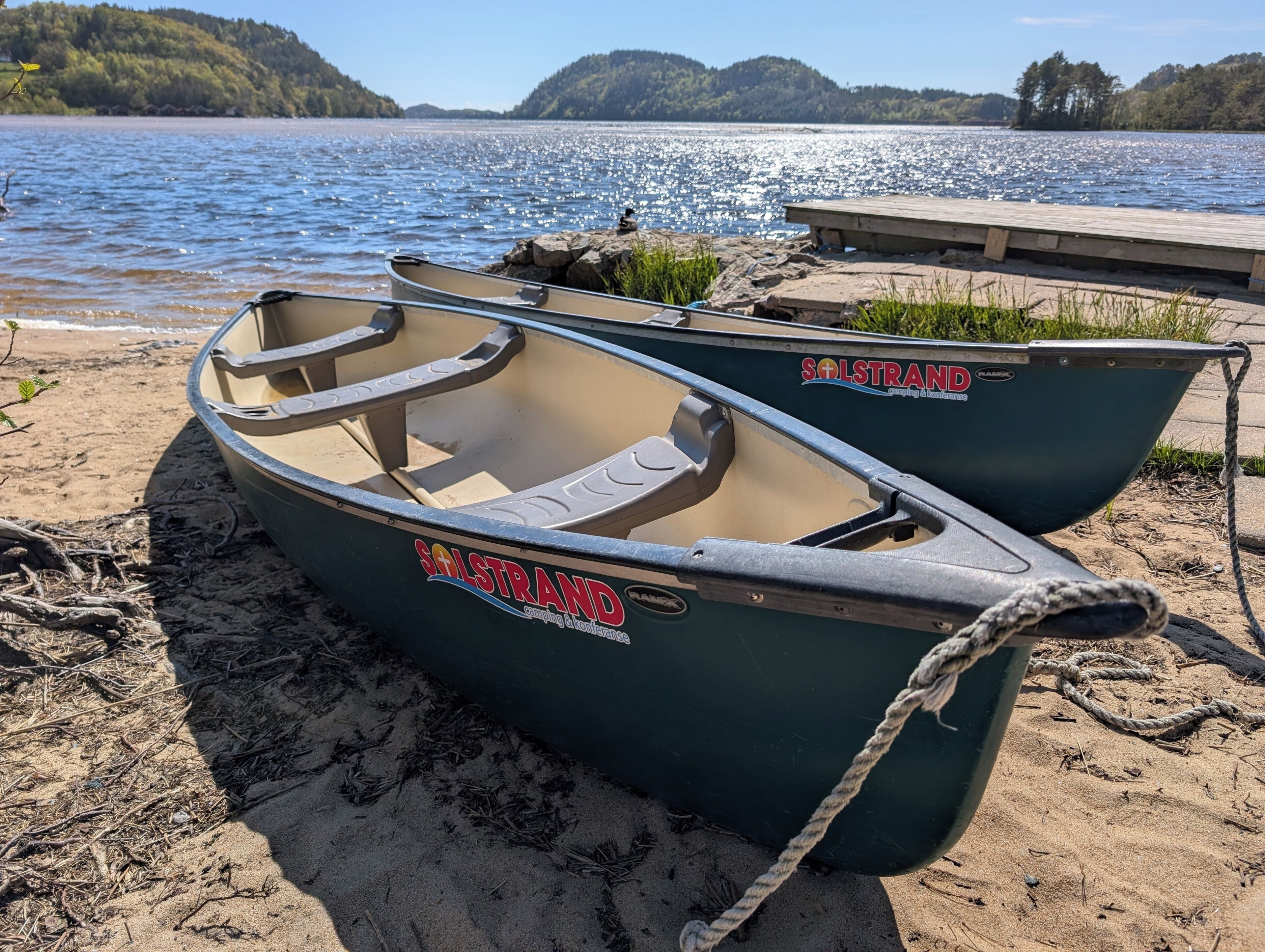 Deux bateaux arborant le logo « Solstrand » sont amarrés sur une plage de sable au bord d’un lac.