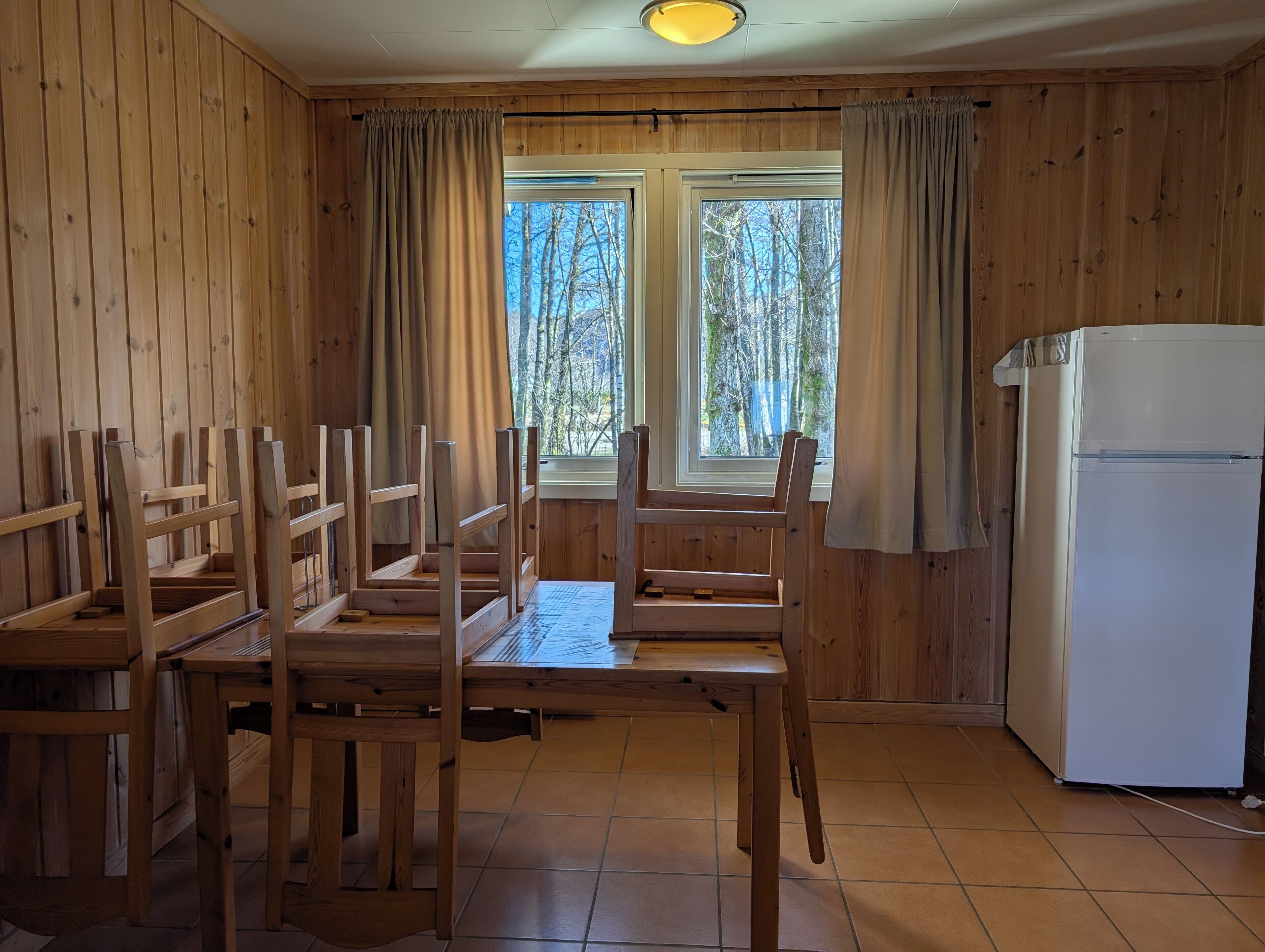 A wooden-paneled room with a dining table and chairs, a refrigerator, and windows with curtains.