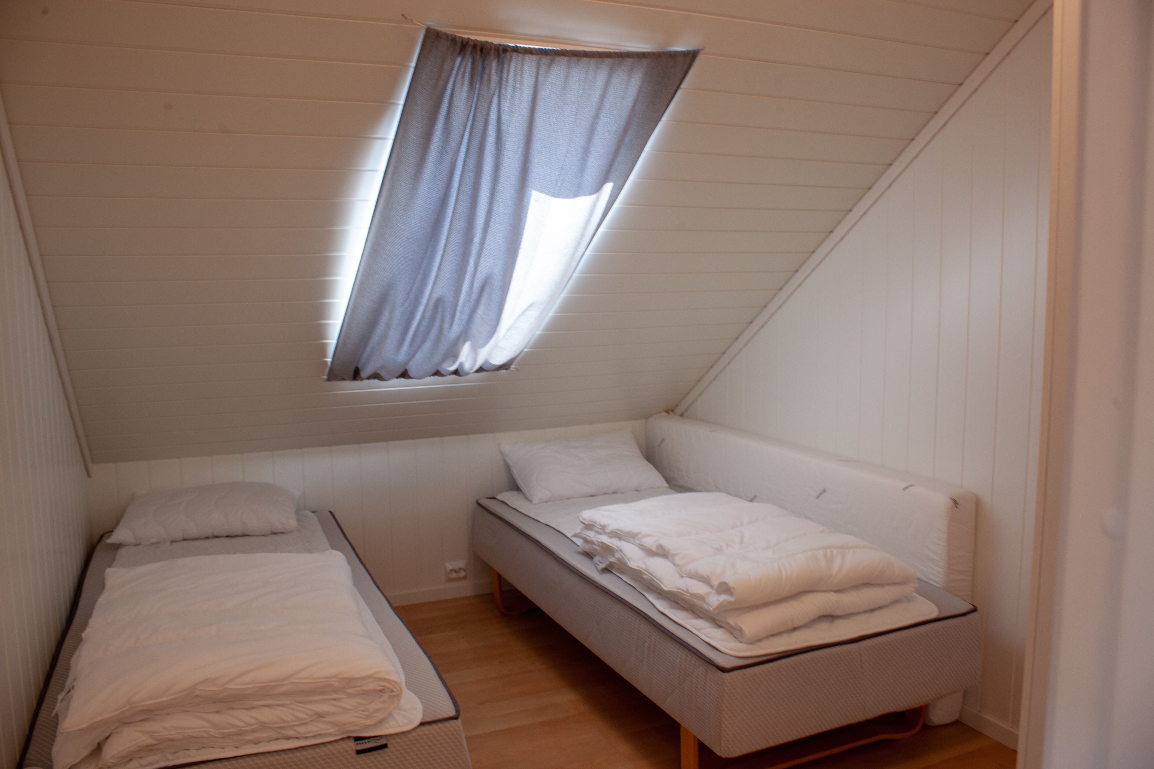 Eine kleine Einzimmerwohnung mit schrägem Dach, einem Fenster mit Vorhang und zwei Betten.