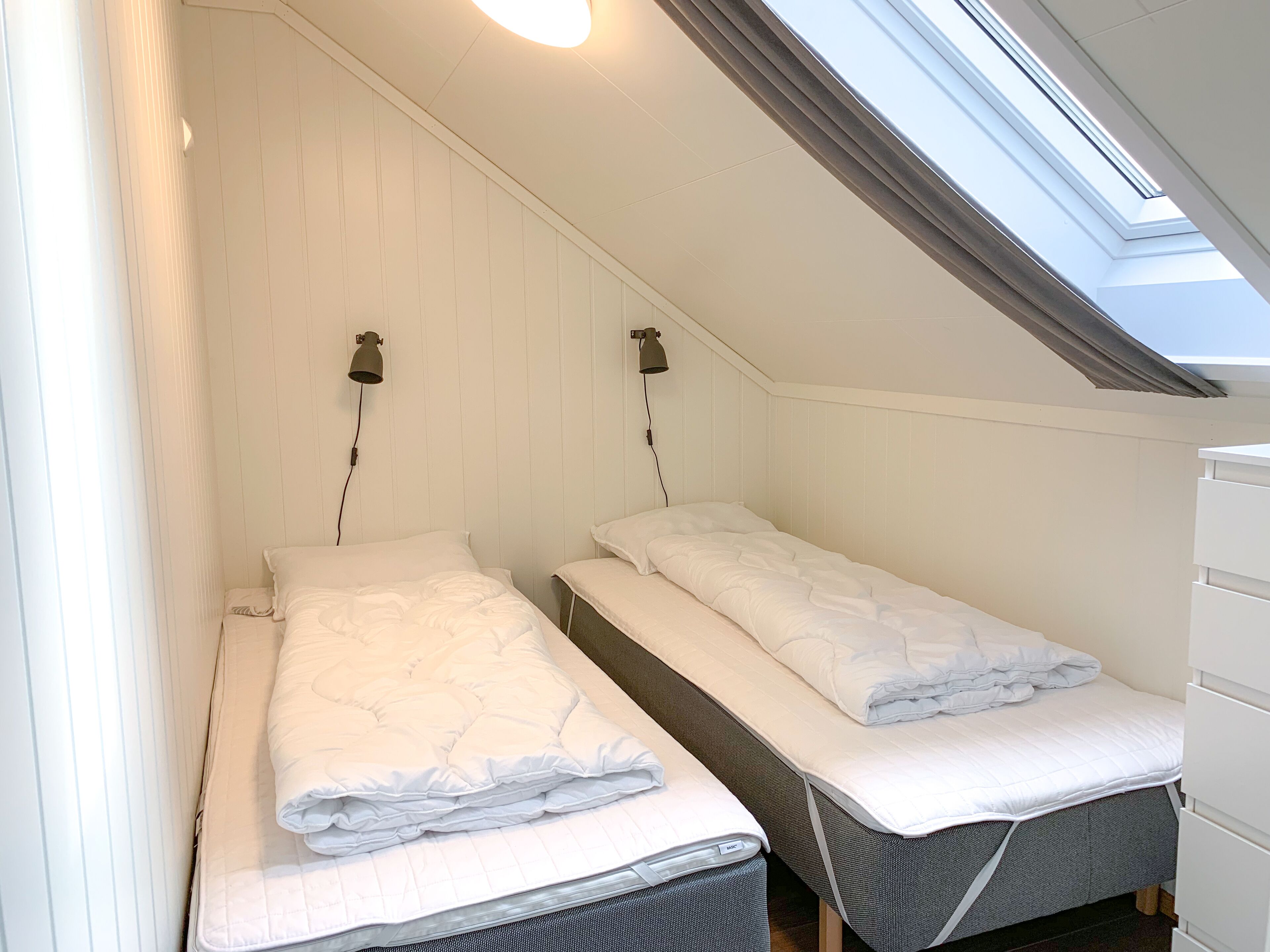 Una habitación pequeña y acogedora con dos camas individuales, un tragaluz y un armario blanco.