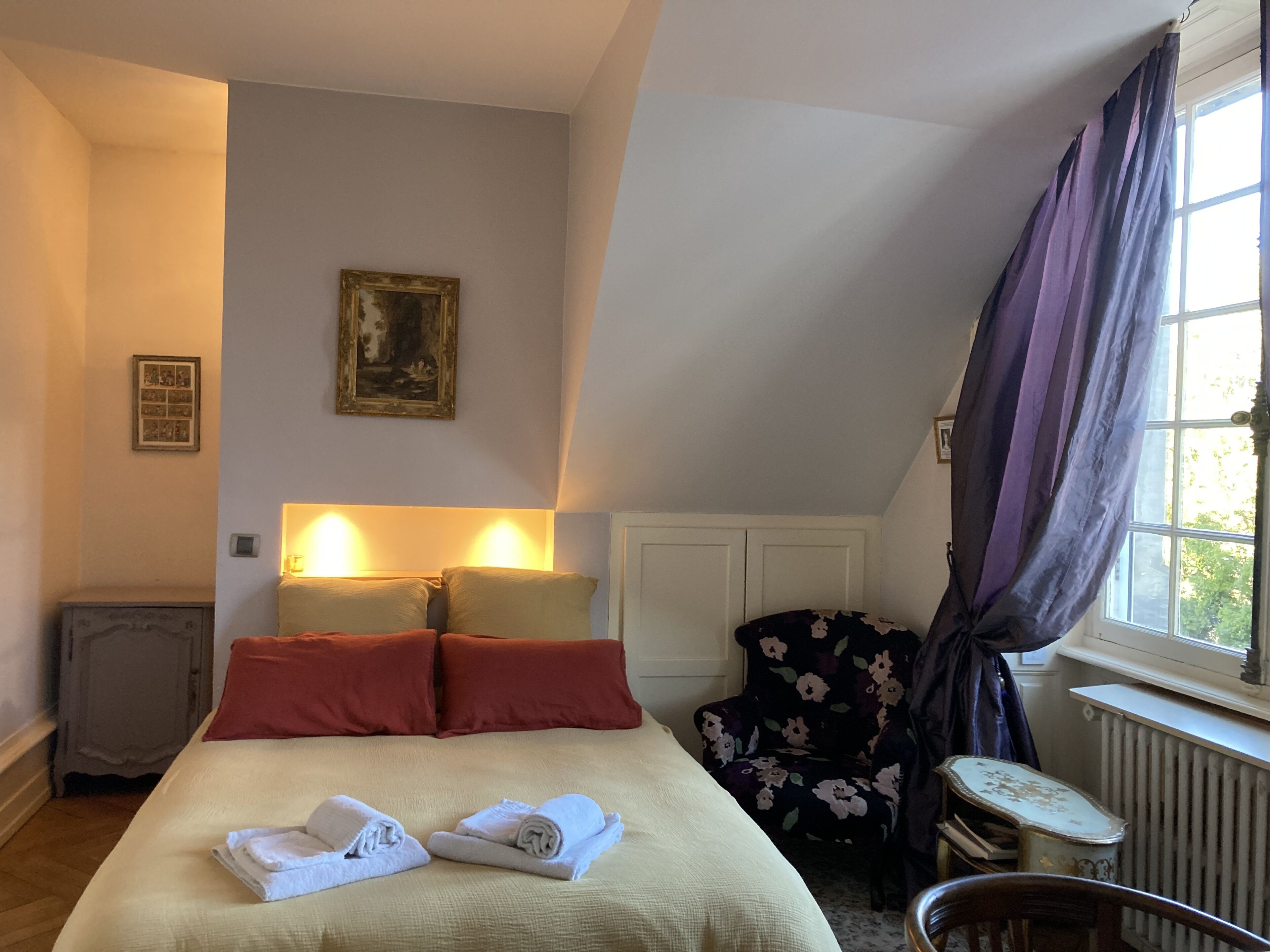 A hotel room with a bed, two bedside lamps, a floral chair, a radiator, and a window with curtains.