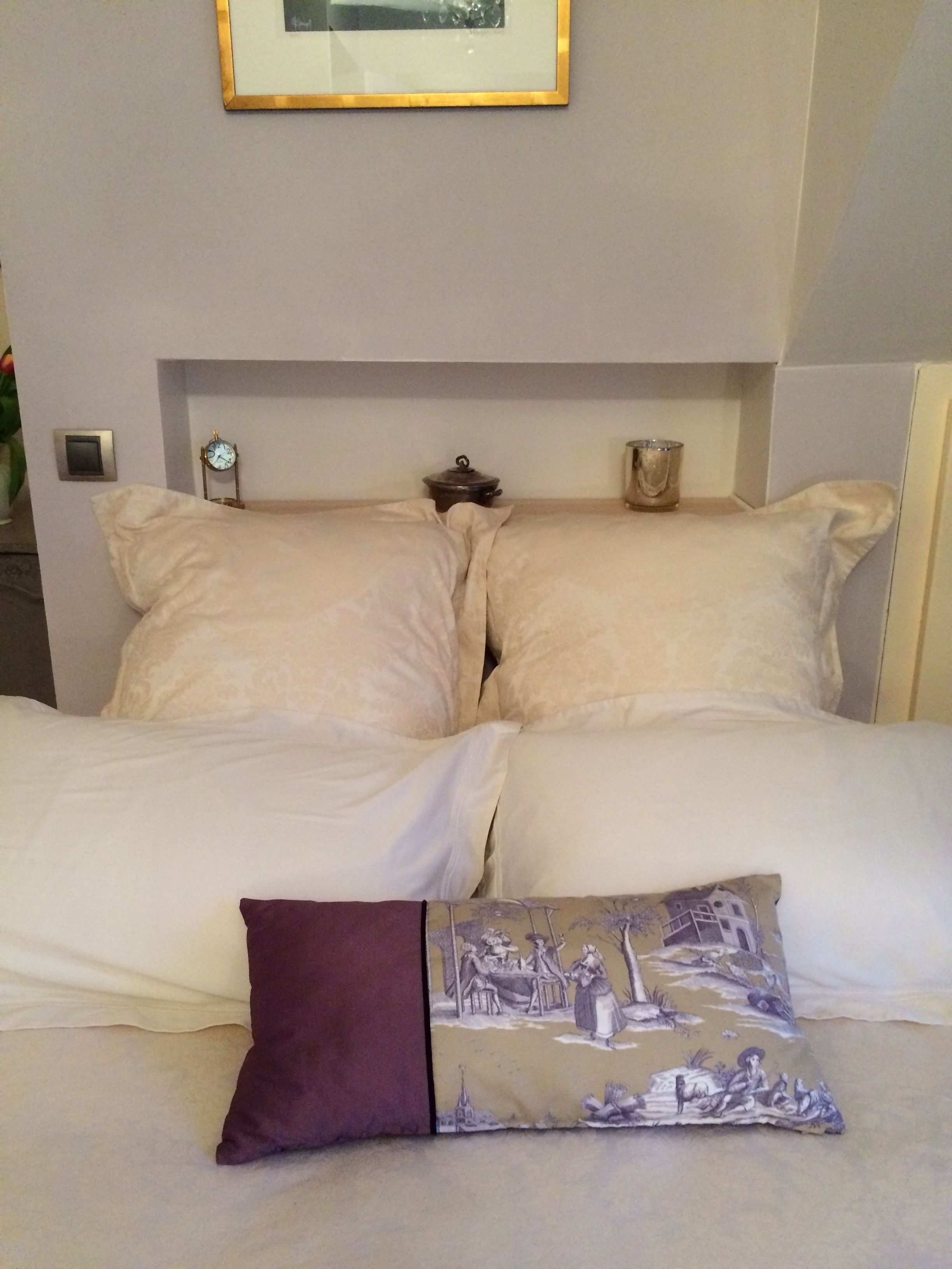 Un lit bien fait, avec deux oreillers et un coussin décoratif, une petite horloge et un tableau encadré accroché au mur.