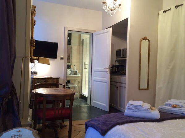 A hotel room with a bed, a desk, a chair, a television, a mirror, and a bathroom visible through an open door. - Chambres d'Hôtes Le Balcon du Prince (Montbeliard)