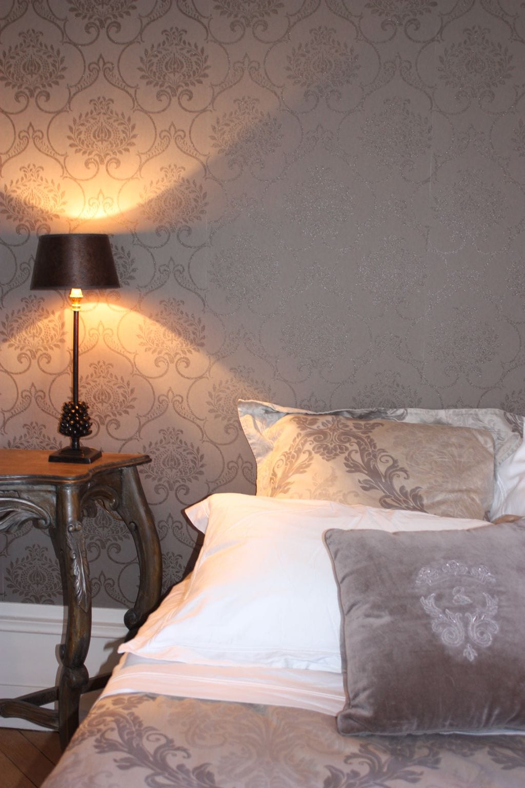 Un dormitorio con papel tapiz estampado, una mesita de noche con lámpara y una cama con almohadas.