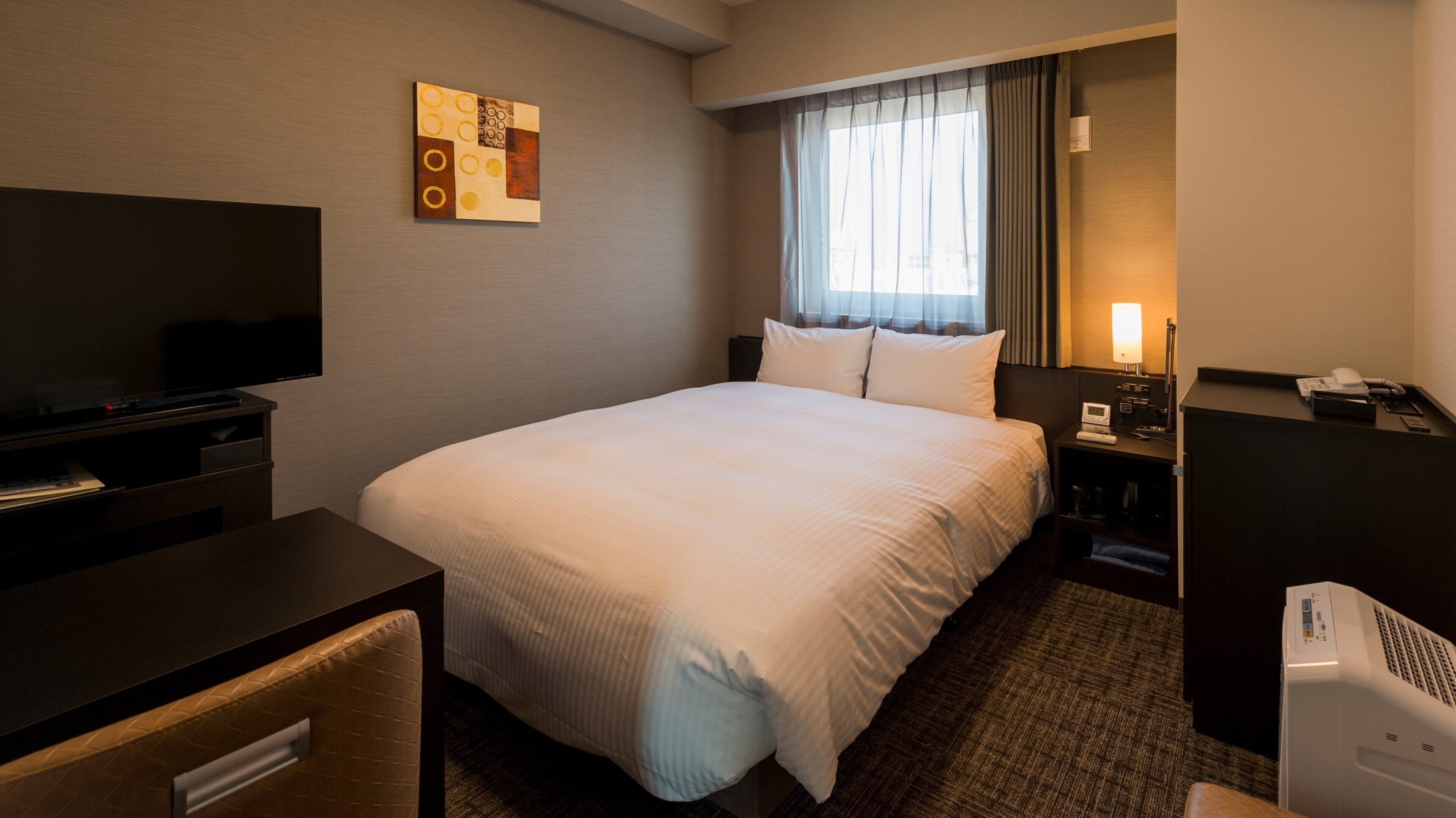 Habitación de hotel con cama, escritorio con televisor, mesita de noche con lámpara y termostato.