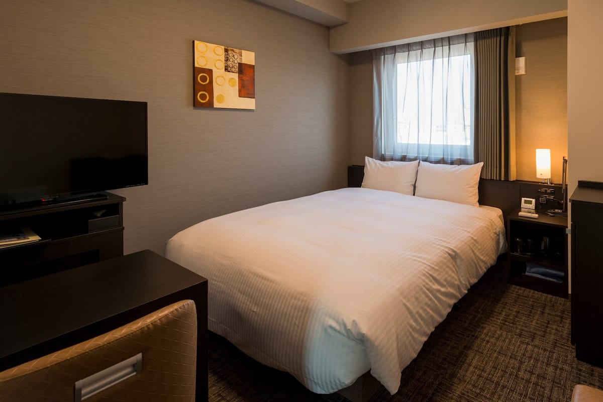 A hotel room with a bed, a desk with a TV, a bedside table with a lamp, and a thermostat.