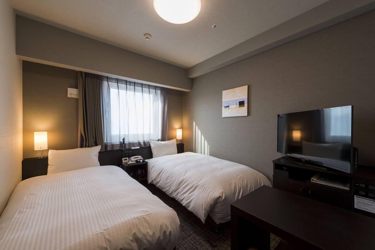 A hotel room with two beds, a TV, a desk, and a window with curtains.