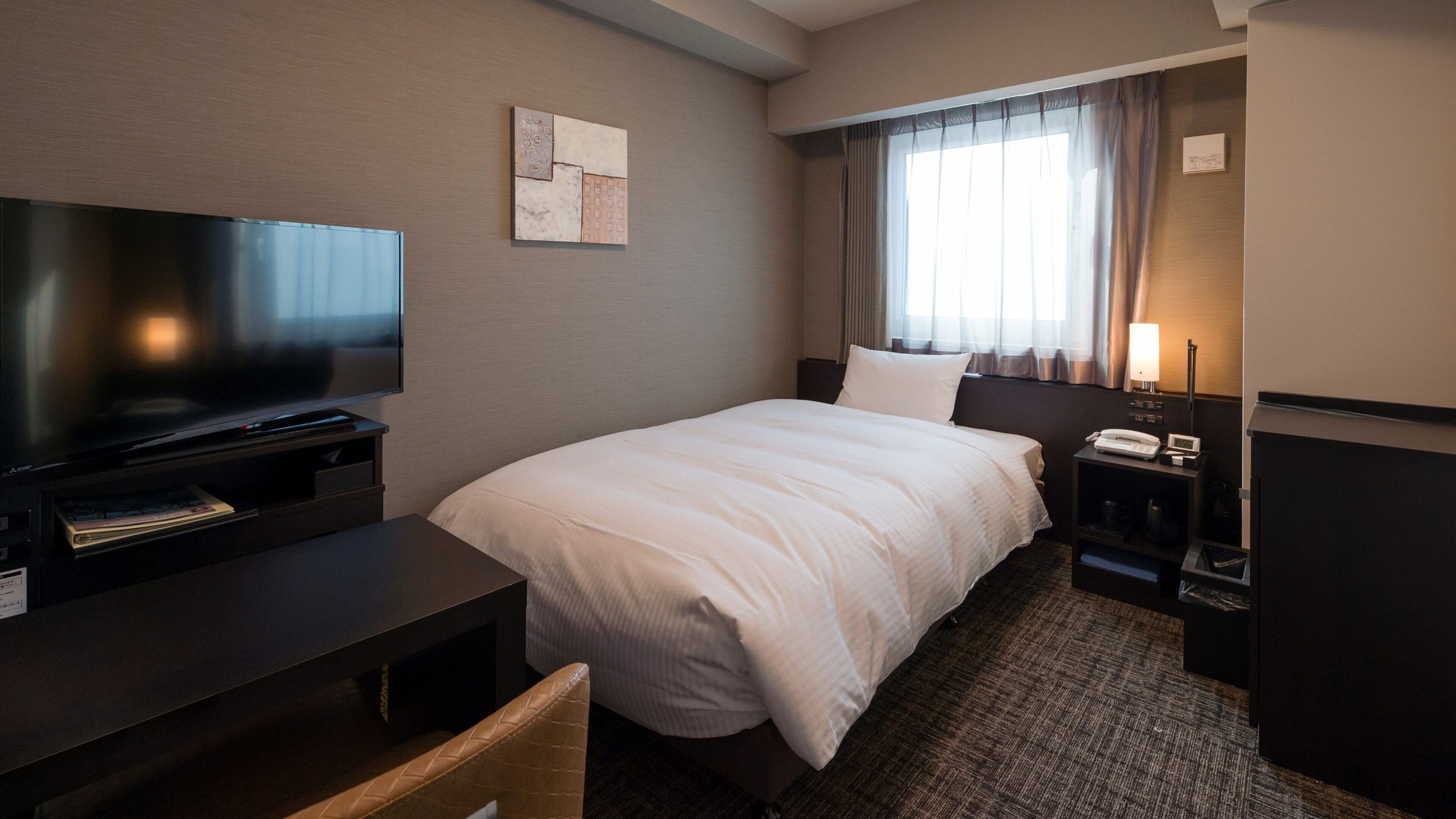 Una habitación de hotel con una cama, televisión, escritorio, lámpara y ventana con cortinas.