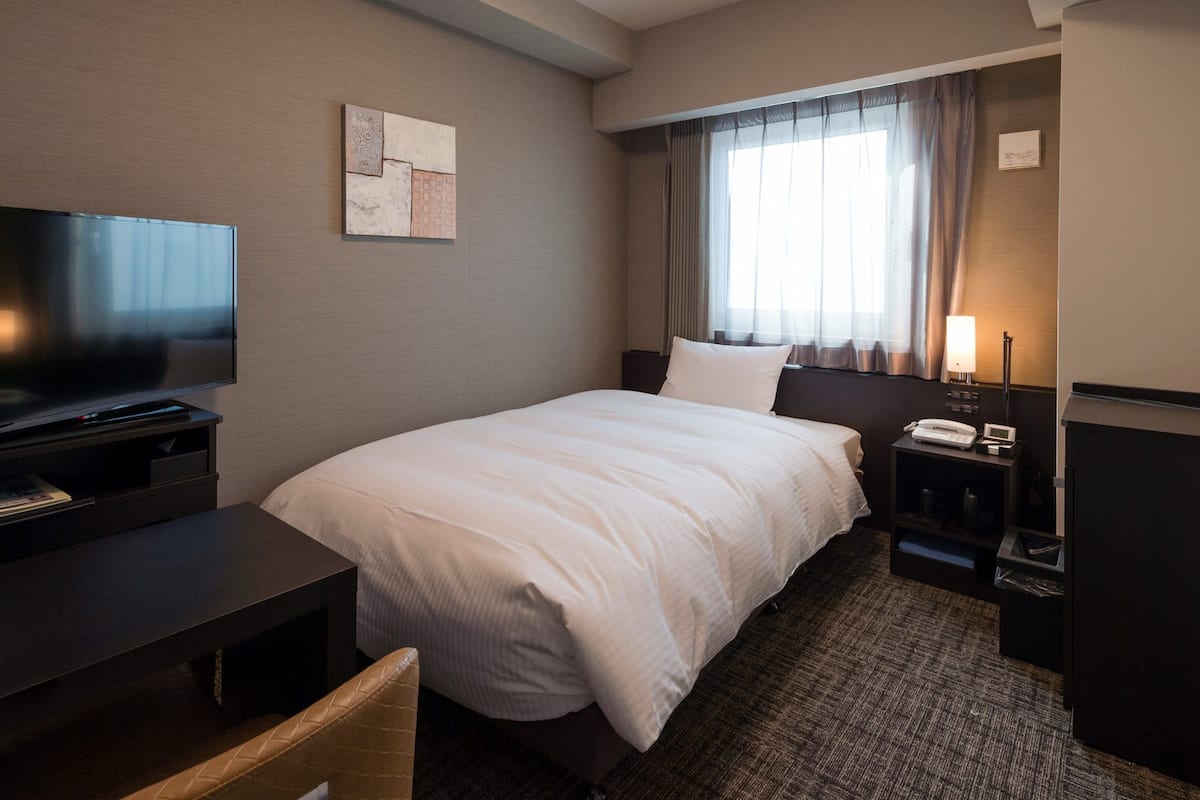 A hotel room with a bed, a TV, a desk, a lamp, and a window with curtains.